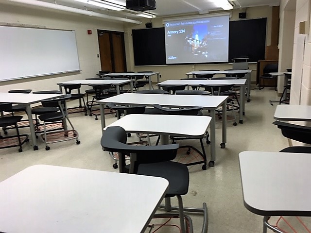 A view of the classroom with variable height, movable desks and chairs; chalkboard; and instructor lectern in front.
