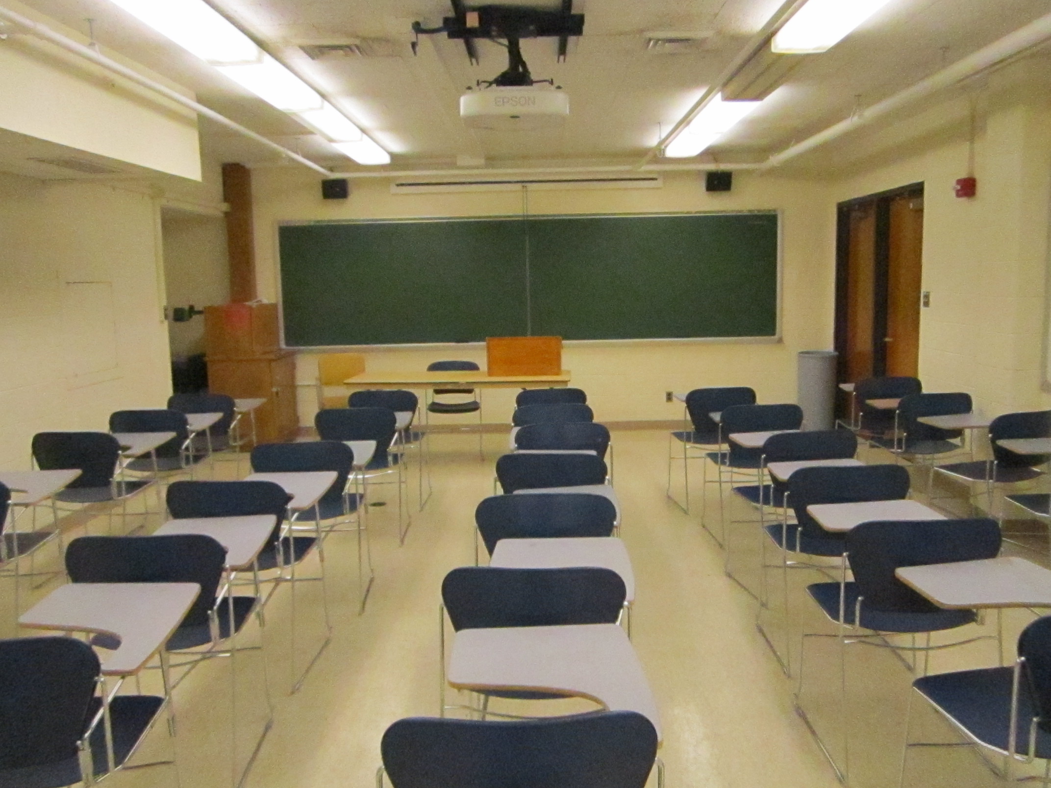 A view of the classroom with movable tableted arm chairs, chalkboard, and instructor table in front