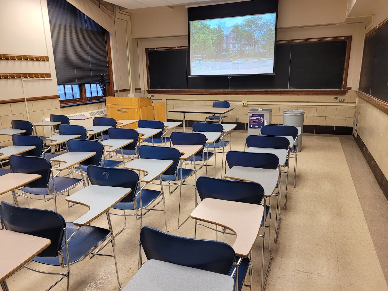 A view of the classroom with movable tablet arm chairs, chalkboard, and instructor table in front.