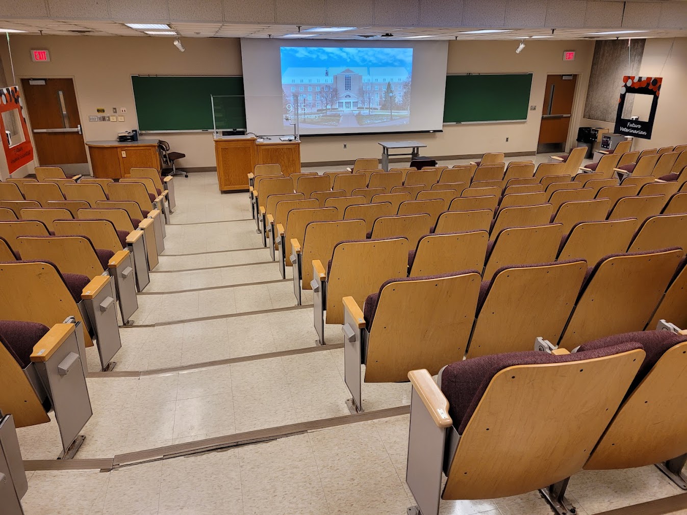 A view of the classroom with theater auditorium seating, chalkboards, lectern, and instructor table in front. 