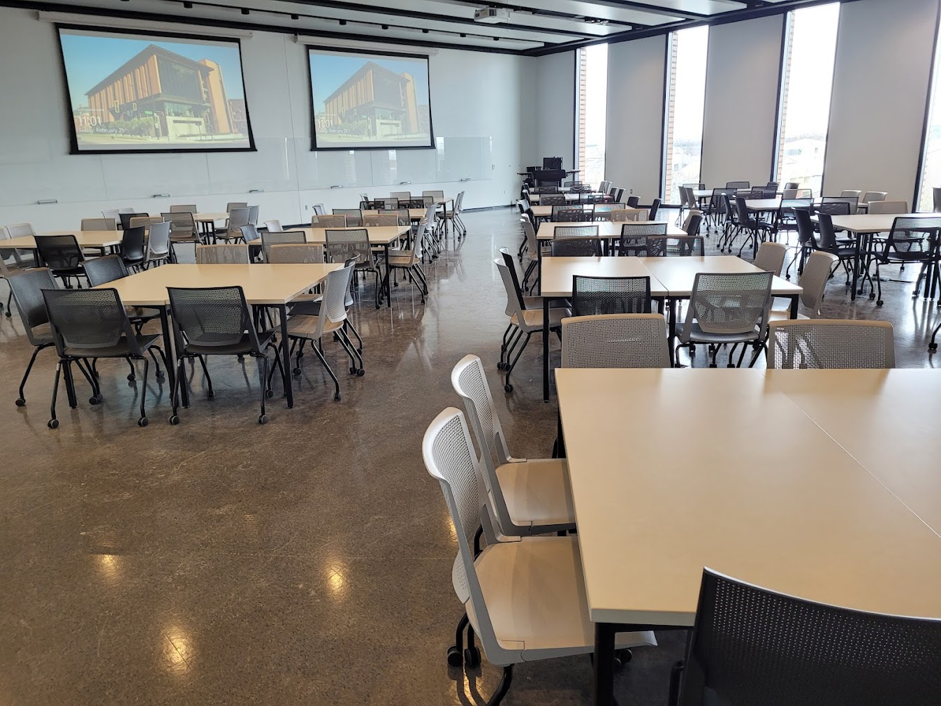 This is a view of the room with student desks, projection screens, a front lecture table, windows, and glass boards.