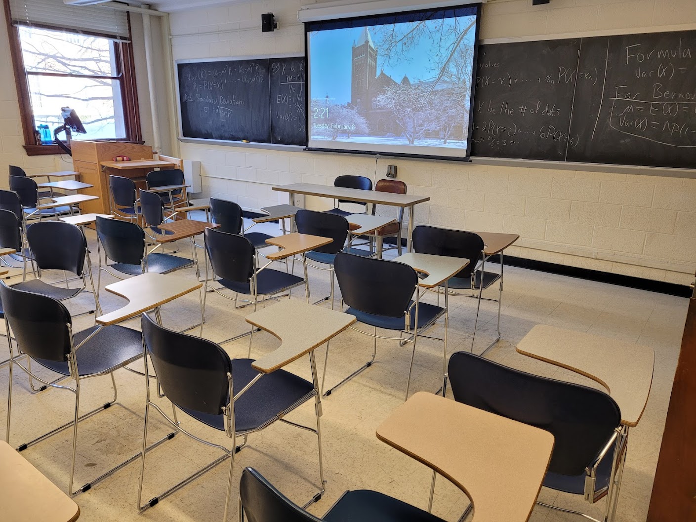 A view of the classroom with movable table arm chairs, chalkboard, and instructor table in front.