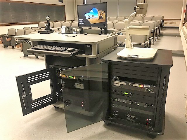 A view of the open cabinet with a computer monitor, keyboard, mouse, microphones, HDMI inputs, a push button controller, document camera, audio visual rack, and electric screen controls.